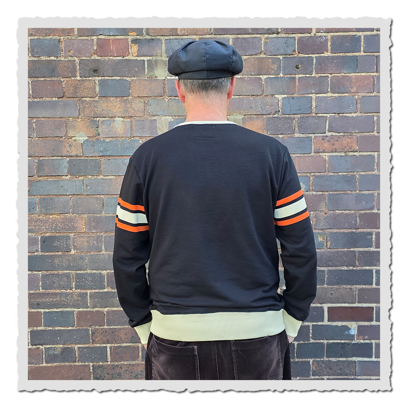 Person von hinten mit schwarzem Sweatshirt und cremefarbenem Saum vor Backsteinwand.